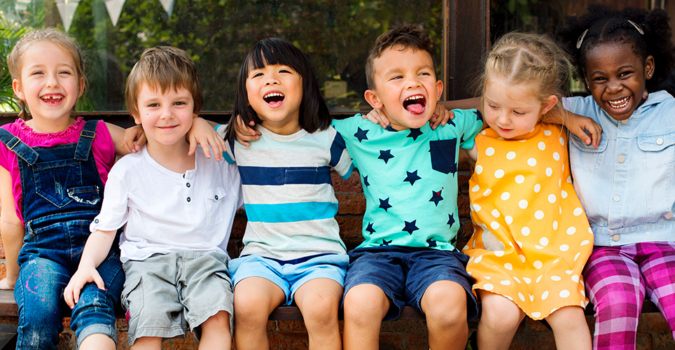 a diverse group of six pre-school kids sitting next to each other, arm in arm with colorful close and big smiles