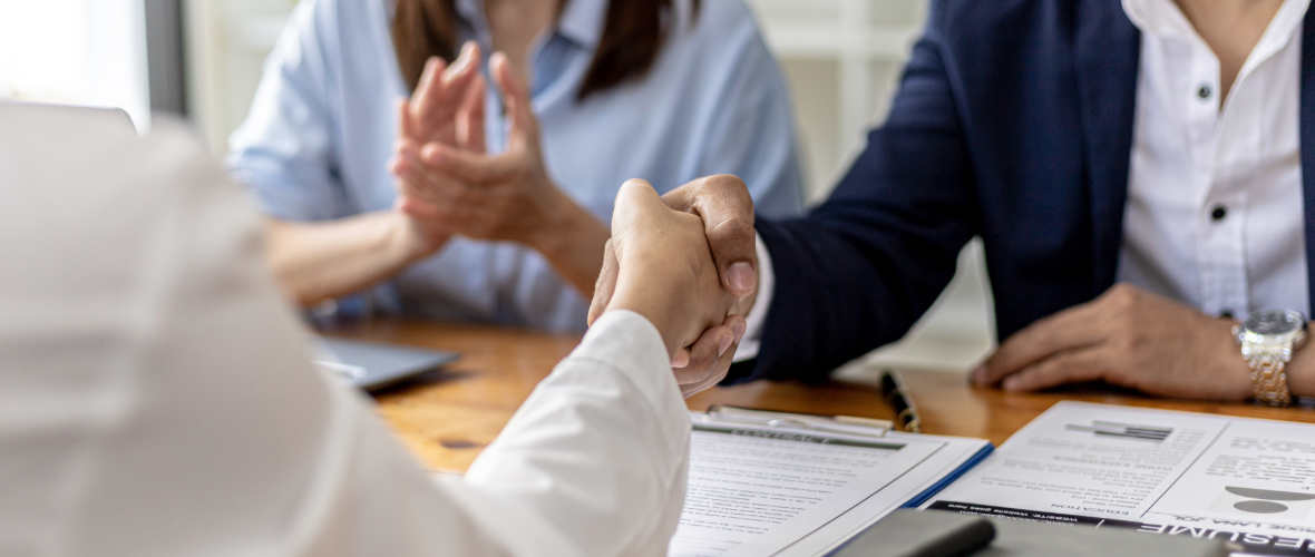 Two people shaking hands across a desk covered with documents.