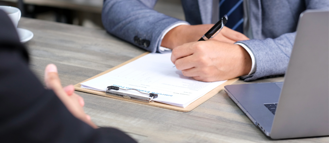 Businessperson filling out a form on a clipboard during an interview, with a laptop on the table.