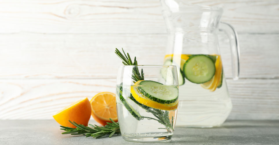 close up of a glass and pitcher on a countertop filled with water, orange slices, cucumber slices and sprigs of thyme
