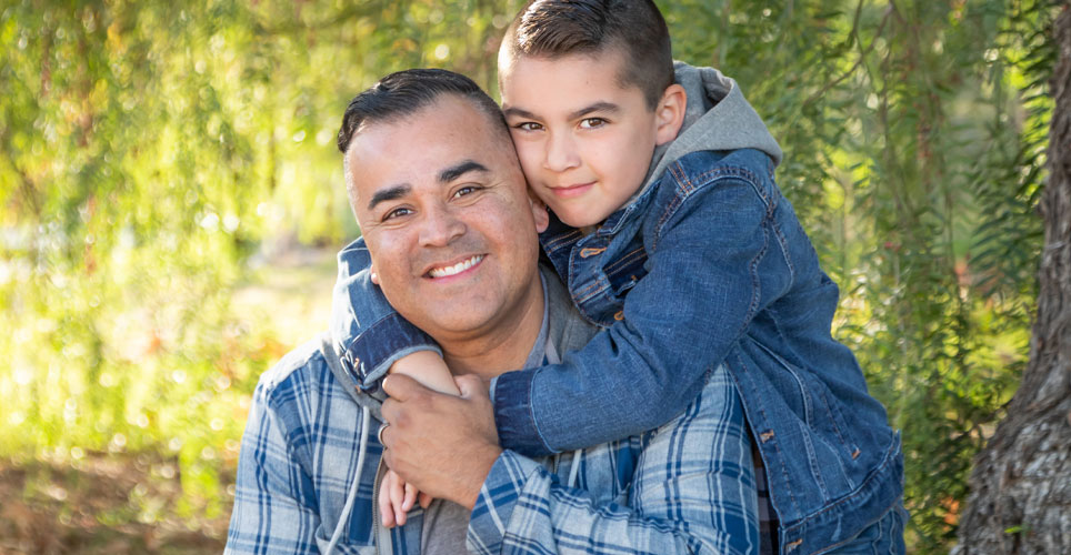 Close up of a Latino father and son, smiling, as the son perches on his father's back.
