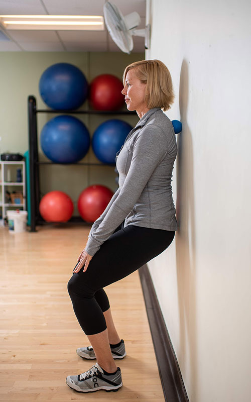 woman standing against wall with a small ball between her upper back and wall showing erector spinae end movement