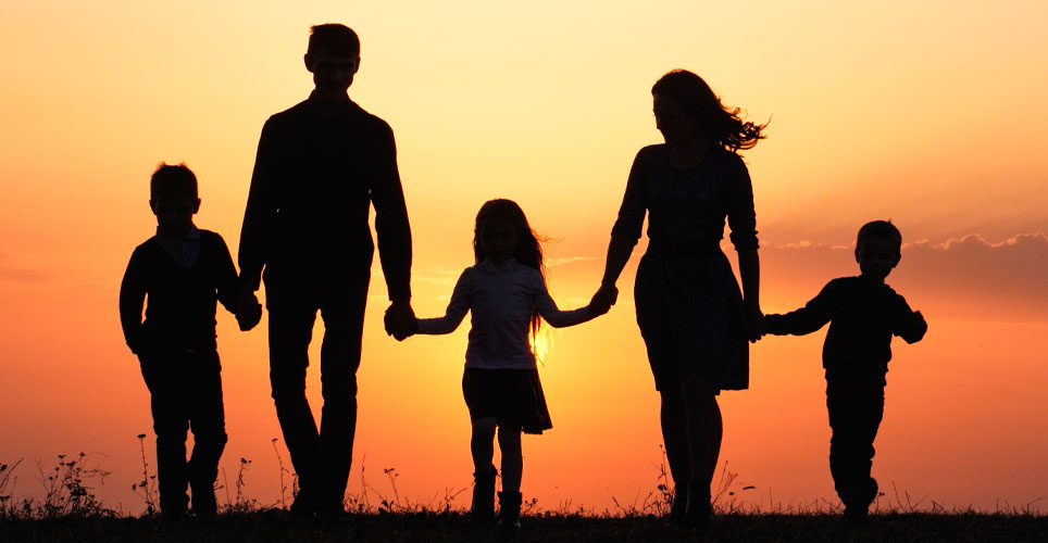 Backlit shot of five people - two adults and three children - holding hands as they walk into a sunset. 