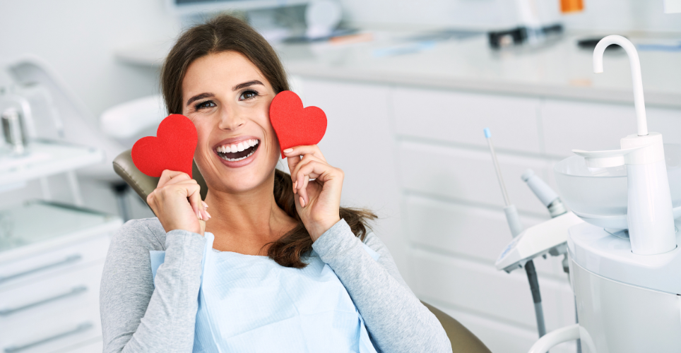 Person sitting in a dental chair holding two red heart shapes over their eyes while smiling, in a dental clinic setting.