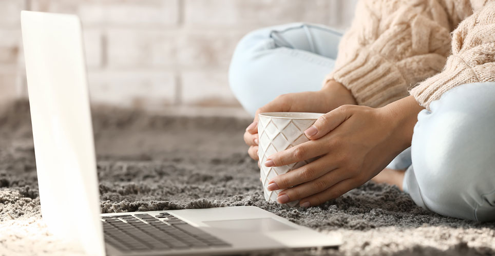 Laptop open on the floor, a woman sitting in front of it with a mug in her hands.