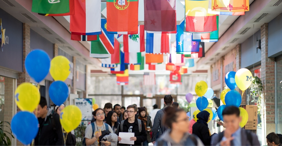 People walking through a hallway with flags from different countries and maize and blue balloons.