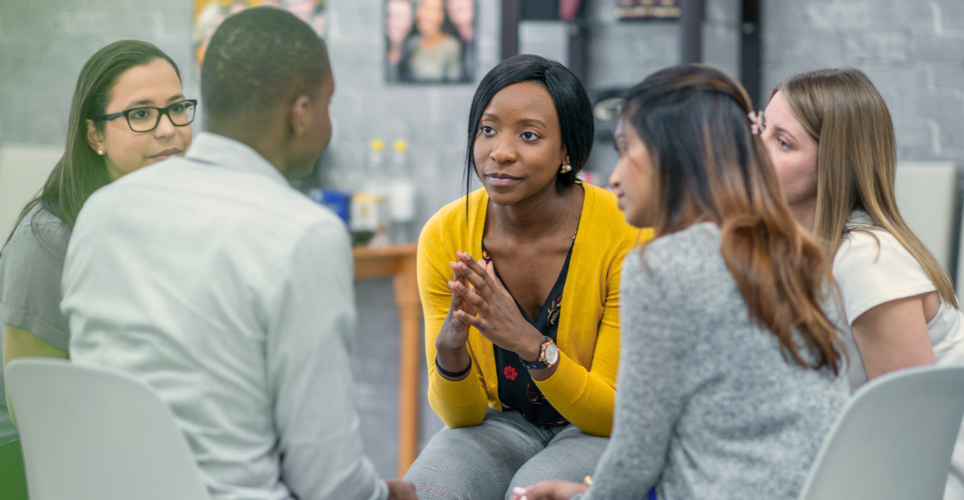 Group of five individuals engaged in a discussion while seated in a casual office setting.