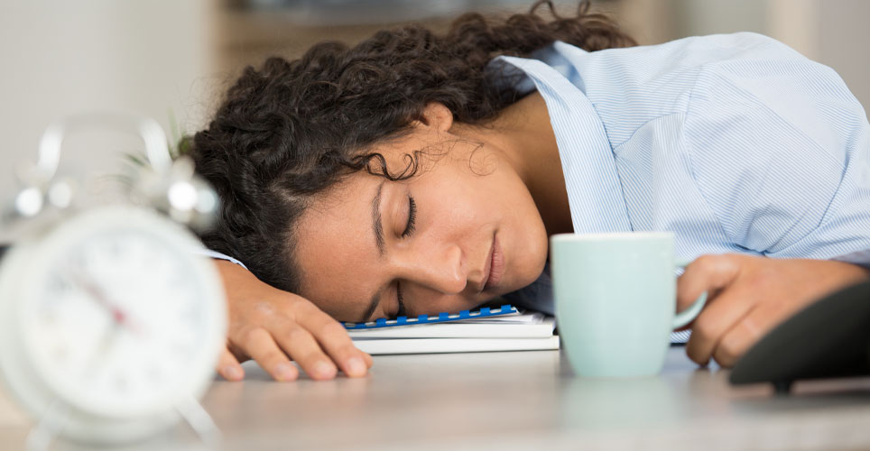 Woman of color sleeping on her desk with close ups of an alarm clock and a coffee cup