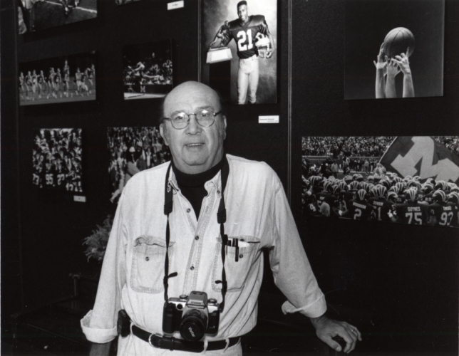 Bob Kalmbach with camera around neck, standing in front of Michigan Football photos