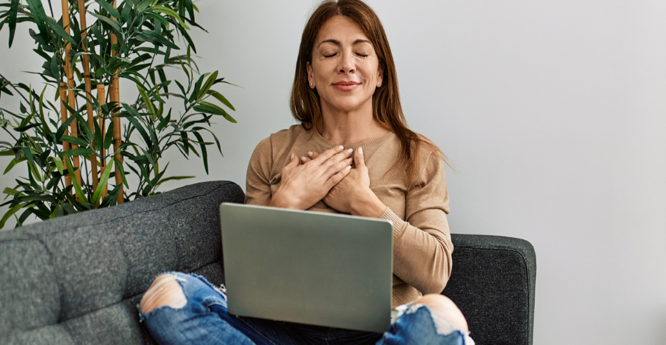woman with brown hair and tan shirt sitting infront of a laptop listening to guided meditation. the woman's eyes are closed and her hands are crossed over her heart.