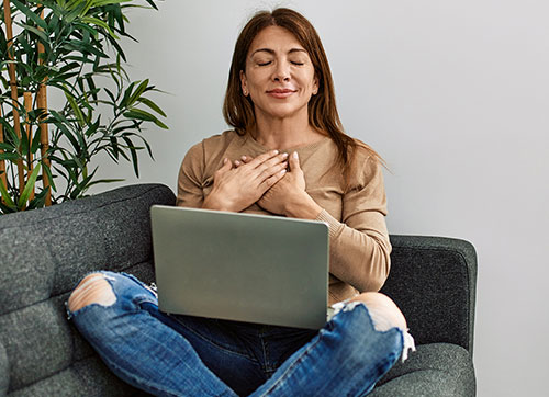 woman sitting on couch, laptop on lap, eyes closed and hands on her chest, meditating