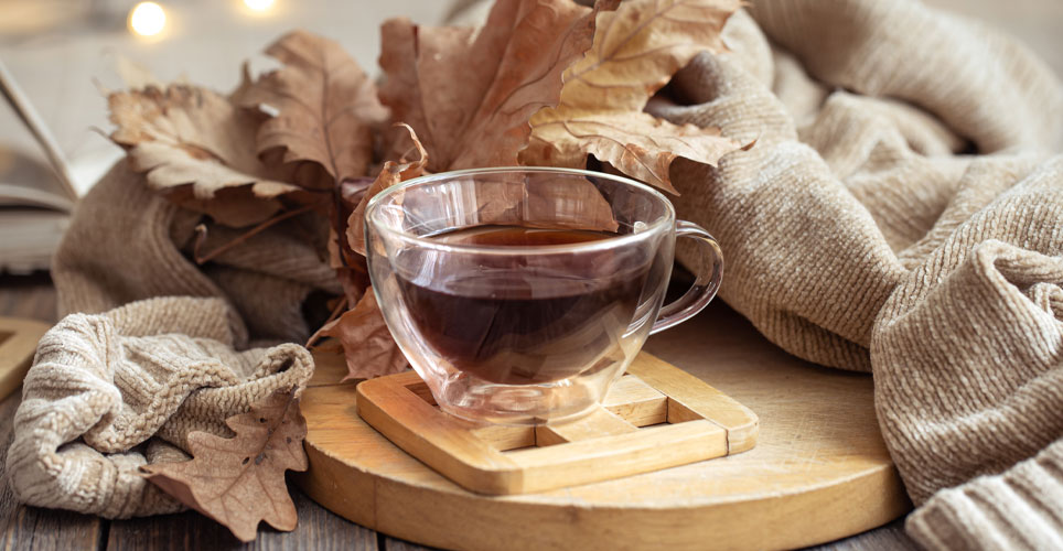 A close up of a transparent cup filled with tea and a tea bag, surrounded by leaves and blankets