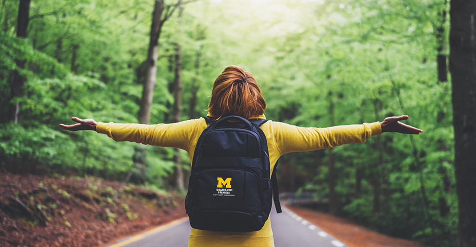 Photo of an African-American woman with her arms spread as she walks with a blue U-M backpack in a green forest