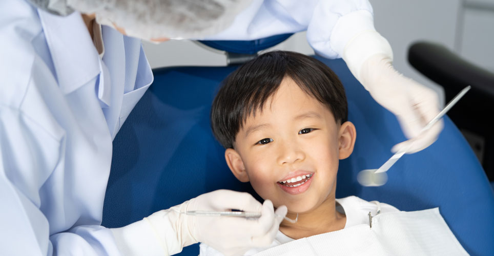 Close up of a small Asian boy in a dentist chair with two gloved hands with instruments near his smiling mouth