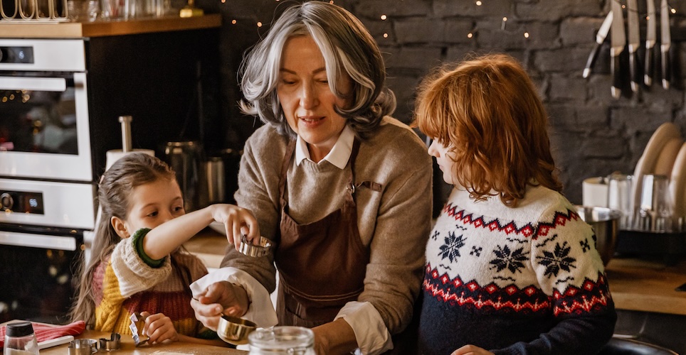 Three people in a kitchen measuring ingredients, gray-haired woman hold a cup with a child on either side looking on.