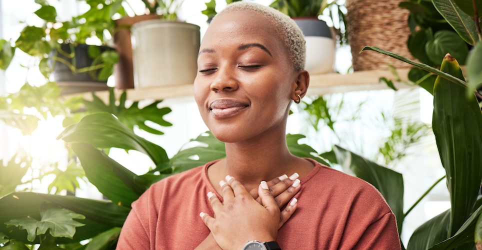 Woman with light brown skin and short hair, standing in a greenhouse with eyes closed and hands crossed over her chest with a calm look on her face.
