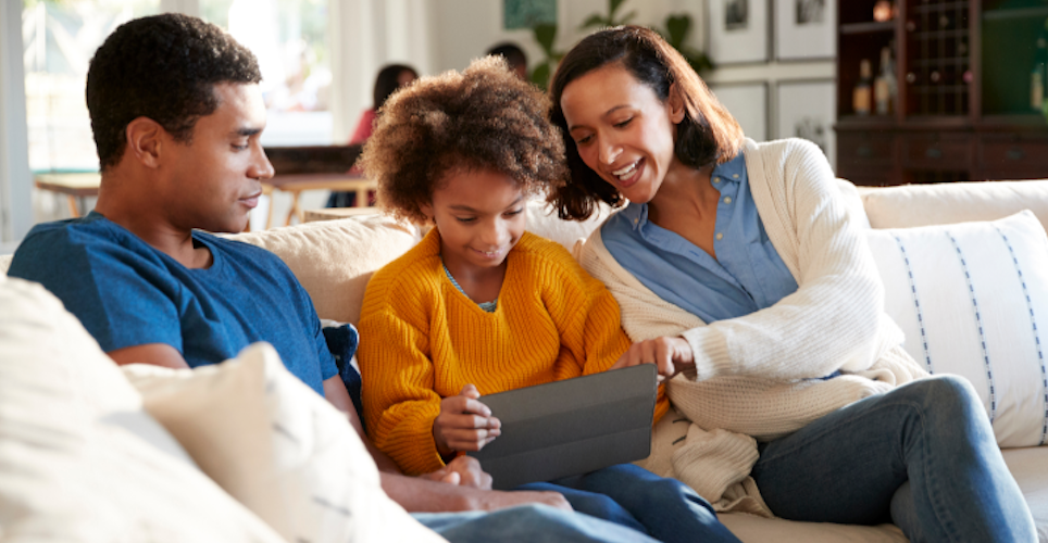African American father, daughter and mother sitting on couch looking at tablet and discussing screen time