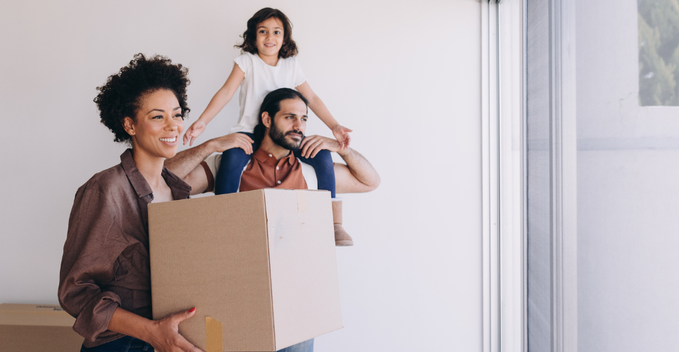 Family of three, with woman holding a moving box and looking out a window. 