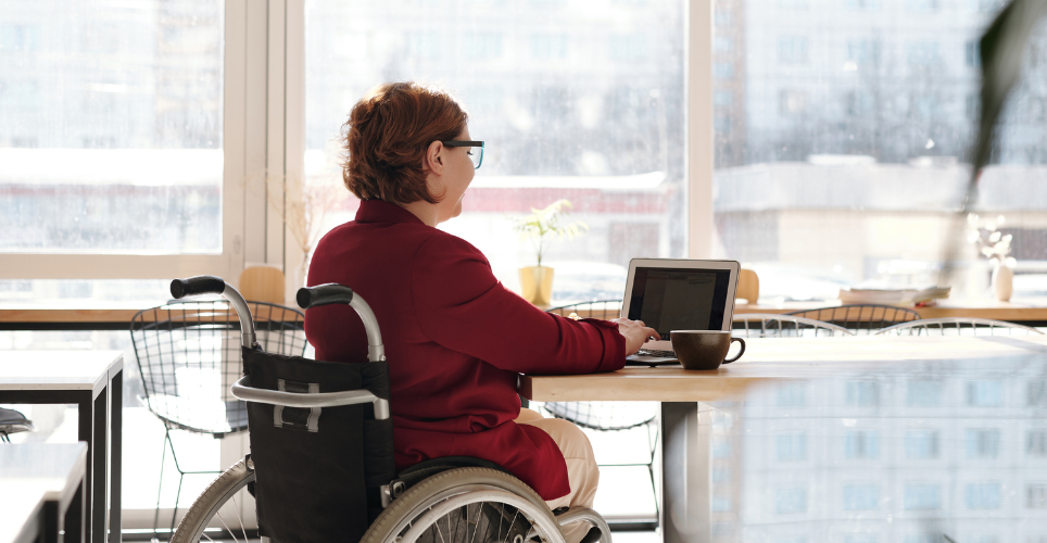 Person in a wheelchair, at a table typing on a laptop with a cup of coffee.