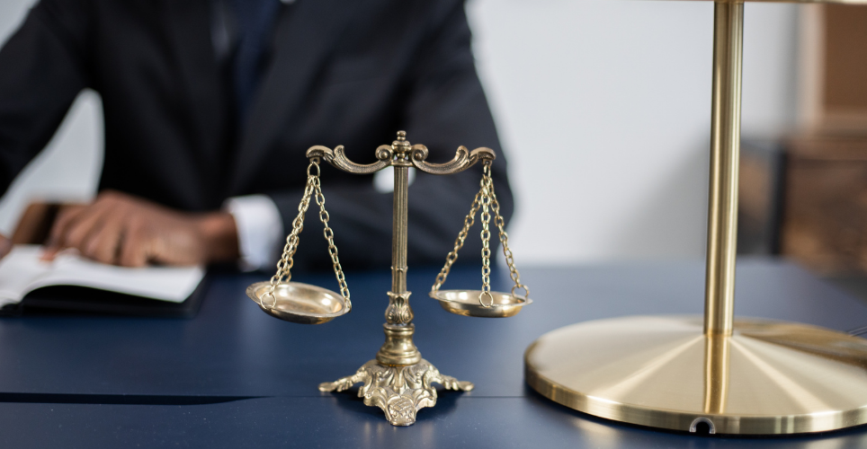 Scales of justice on a desk in front of a man sitting at the desk with his hands resting on paperwork