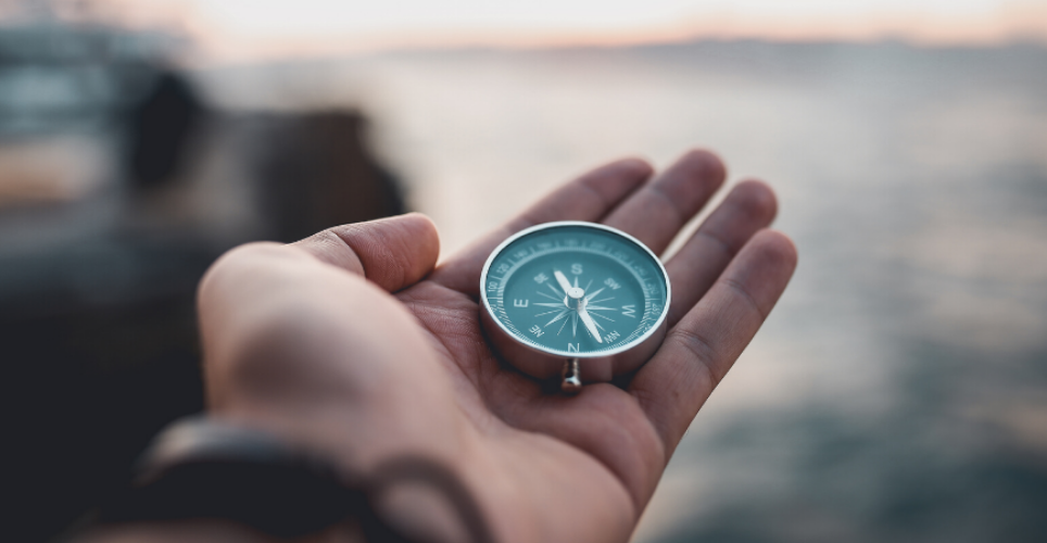 Light skinned hand holding a compass with a natural water scene blurred in the background.