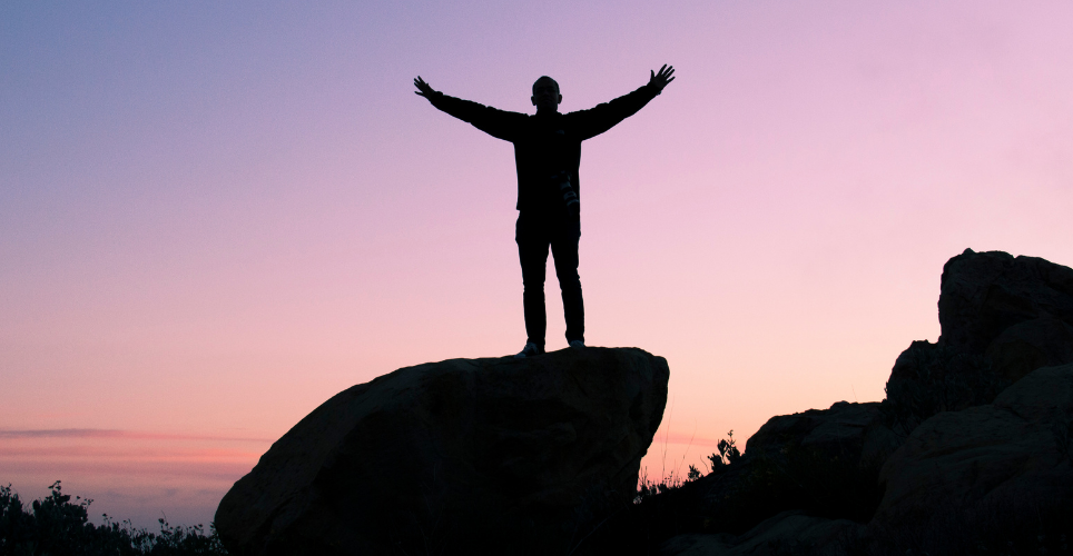 silhouette of person with arm wide, standing on a rock, looking into the sunset 