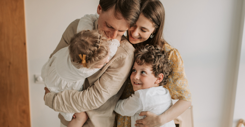 Family embracing, A man holding a toddler with a young child hugging his waist and looking up smiling. a woman hugging all three smiling.