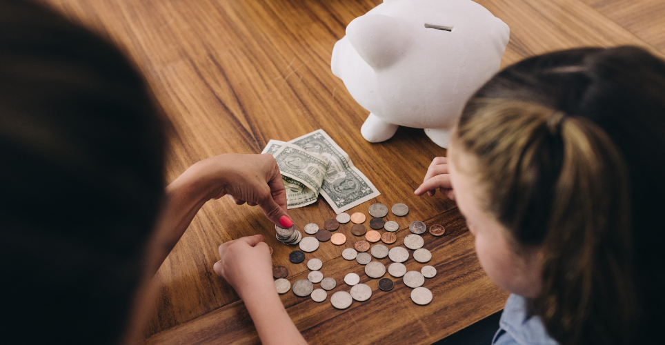 Young girl and her mom counting money on a table next to a piggy bank.