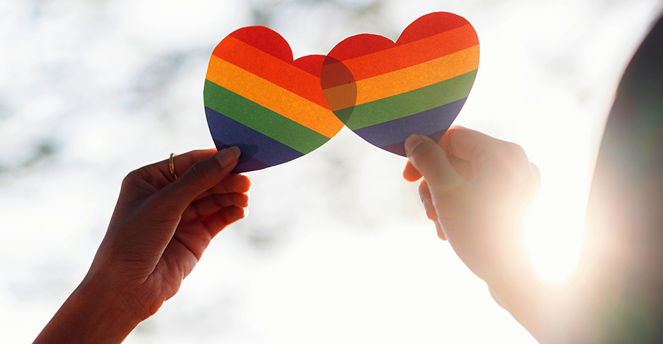close up two hands holding rainbow cutout hearts that are touching