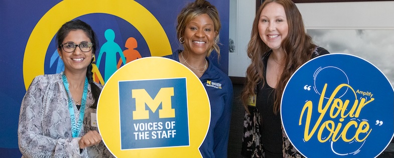 Three voices team members, smiling, and holding photo props with "Voices" logo standing in front od a colorful background.