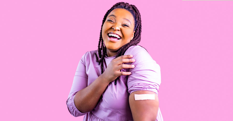 photo of woman of color smiling and showing a bandaid (indicating she received a vaccination) on a bright pink background